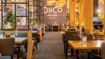 Interno di un ristorante con diversi tavoli e sedie, una parete ampia con il nome 'DUCO HELMOND by Fletcher' e piante pensili.