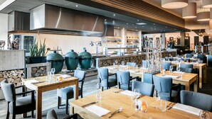 Un restaurante moderno con mesas de madera y sillas azules, una cocina al fondo y jarrones decorativos sobre las mesas.