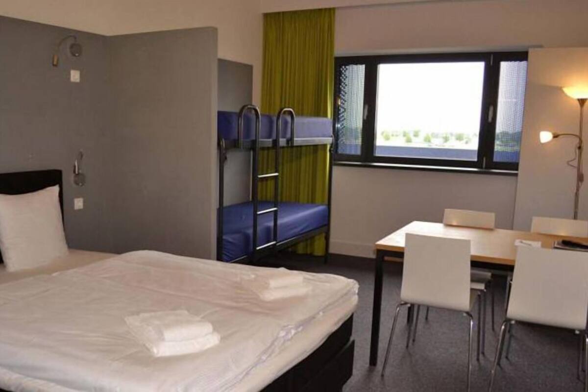 A hotel room with a single bed, a desk with two chairs, a window with a view, and a bunk bed with blue bedding.