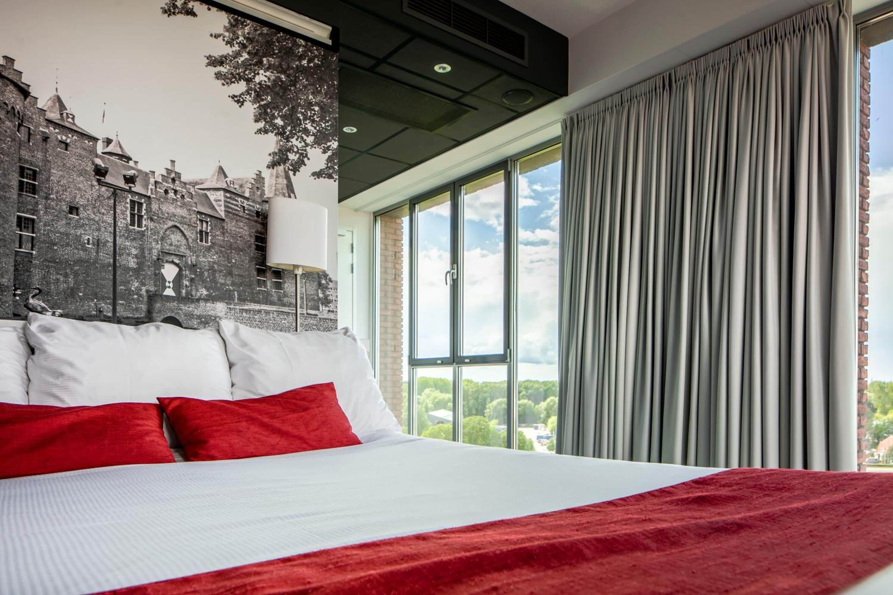 A hotel room with a large bed, red and white bedding, a mural of a castle on the wall, and a view of greenery through large windows.