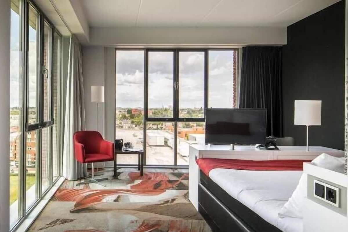 A hotel room with a bed, a desk, a chair, a TV, and large windows offering a city view.