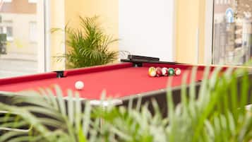 Une table de billard avec des billes et une queue, entourée de plantes vertes.