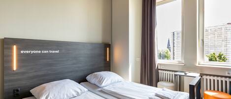 Una habitación de hotel con una cama, una ventana con cortinas, un radiador y un taburete naranja pequeño.