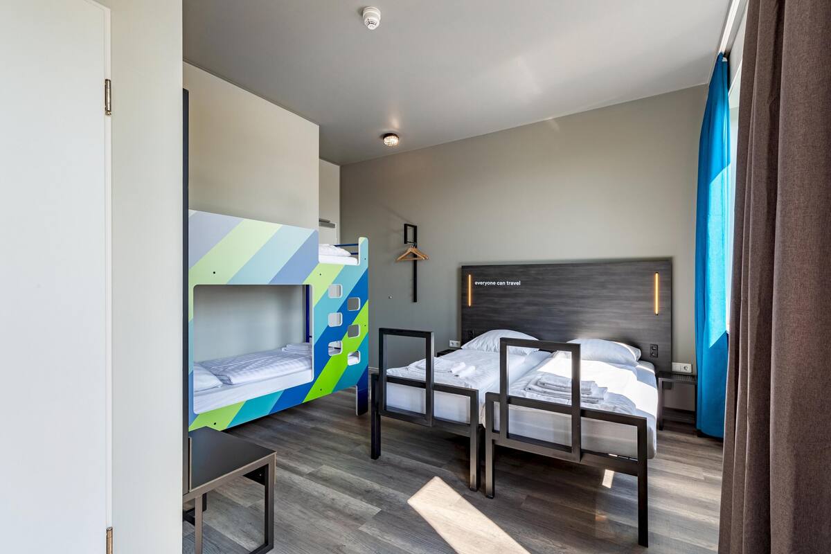 Habitación de hotel con literas, una mesita de noche, un perchero y un cabecero con lámpara.