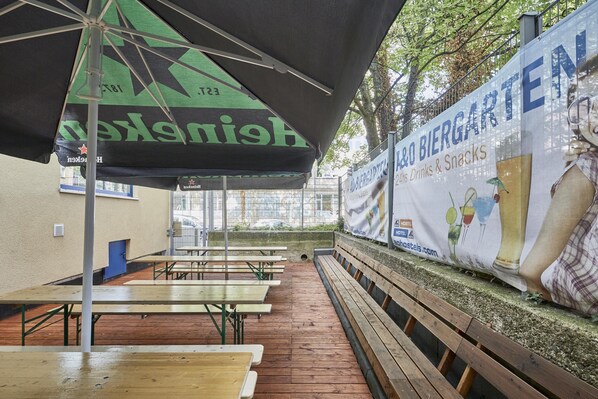 An outdoor seating area with tables and benches, a green and black umbrella, and a banner advertising a beer garden. - a&o Aachen Hauptbahnhof - Hostel (Aachen)