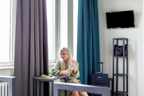 A hotel room with a bed, a table with a bottle and snacks, a suitcase, a TV on the wall, and curtains. - a&o Aachen Hauptbahnhof - Hostel (Aachen)