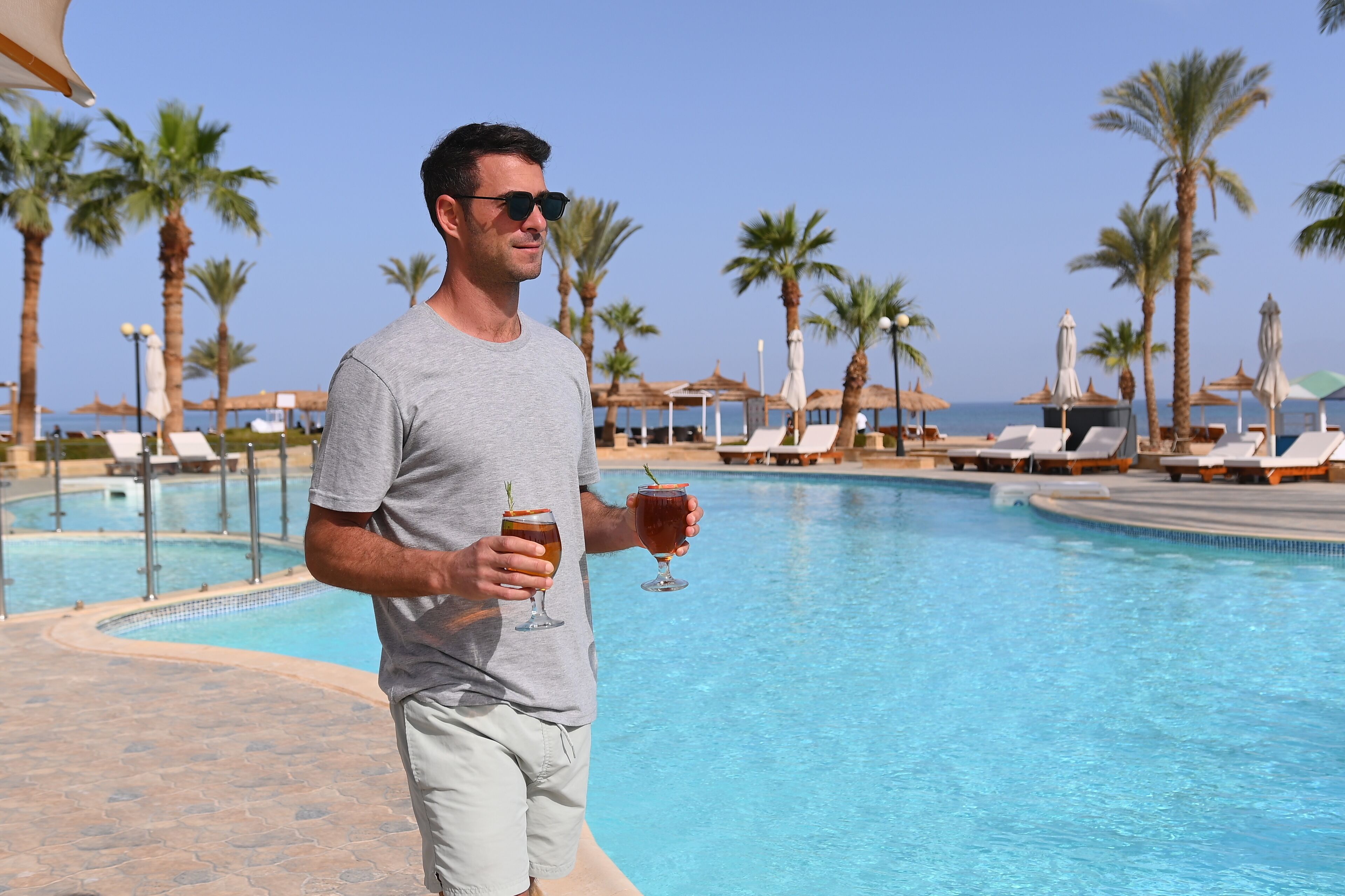 Un homme se tient près d’une piscine, avec des chaises longues et des palmiers en arrière-plan.