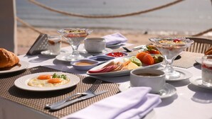 Ein Tisch ist reichhaltig mit Frühstücksleckereien wie Eiern, Obst und Gebäck gedeckt, im Hintergrund ist der Blick aufs Meer.