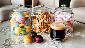 A glass table with three glass jars containing colorful candies, waffles, and marshmallows, and a cup of coffee. - Hotel Het Heilig Genot (Vleteren)