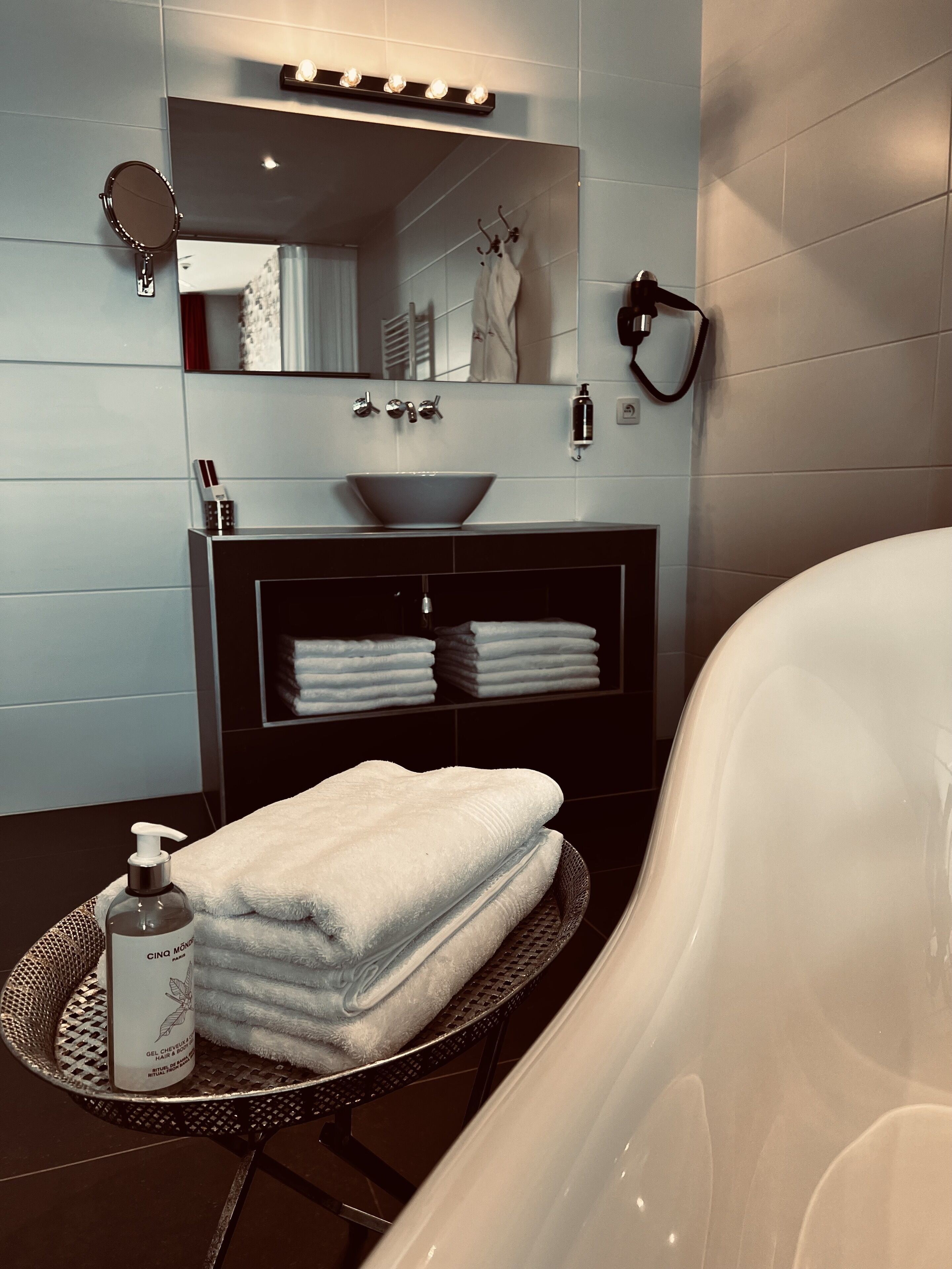 Ein modernes Badezimmer mit einer großzügigen Badewanne, einem runden Waschbecken mit einer Haube, einem Spiegel und einem Handtuchhalter. Auf einem runden Tablett liegen gefaltete Handtücher und am Waschbecken steht eine Flasche Handseife.