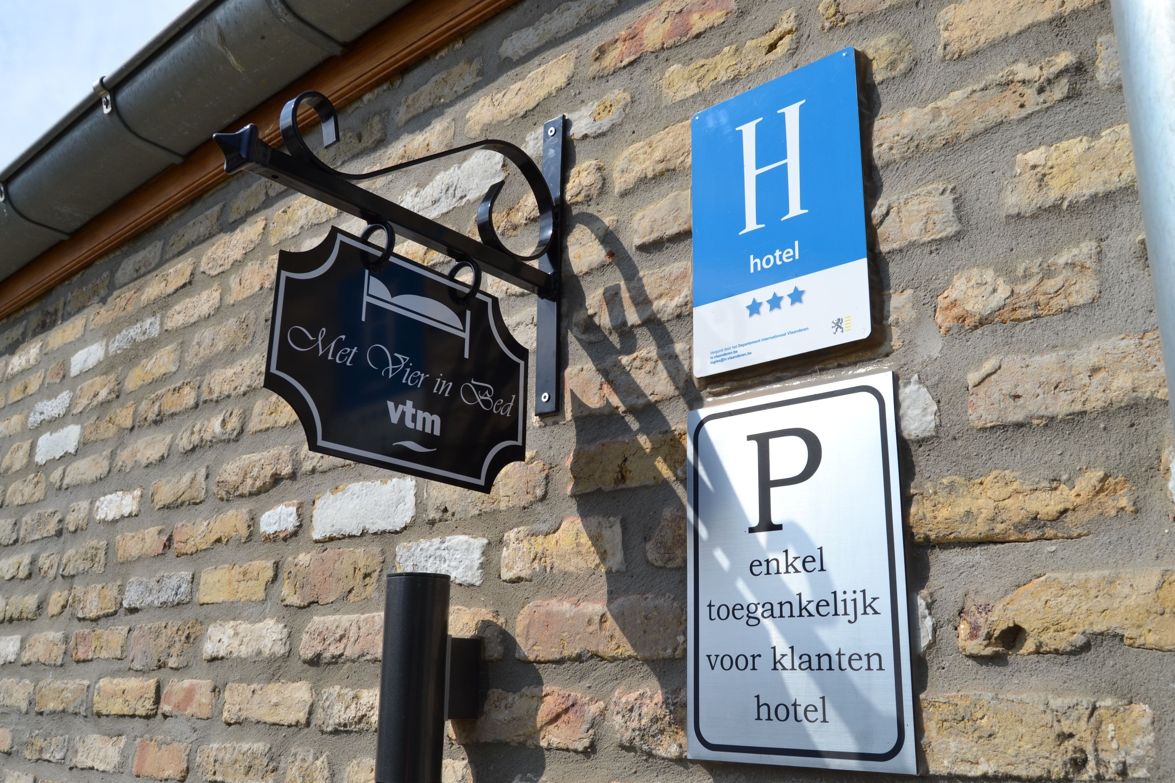 A hotel sign with a blue background and a white 'H' for hotel, a black sign with 'Met Jier in God' and 'Vtm', and a white sign with a black 'P' for parking.