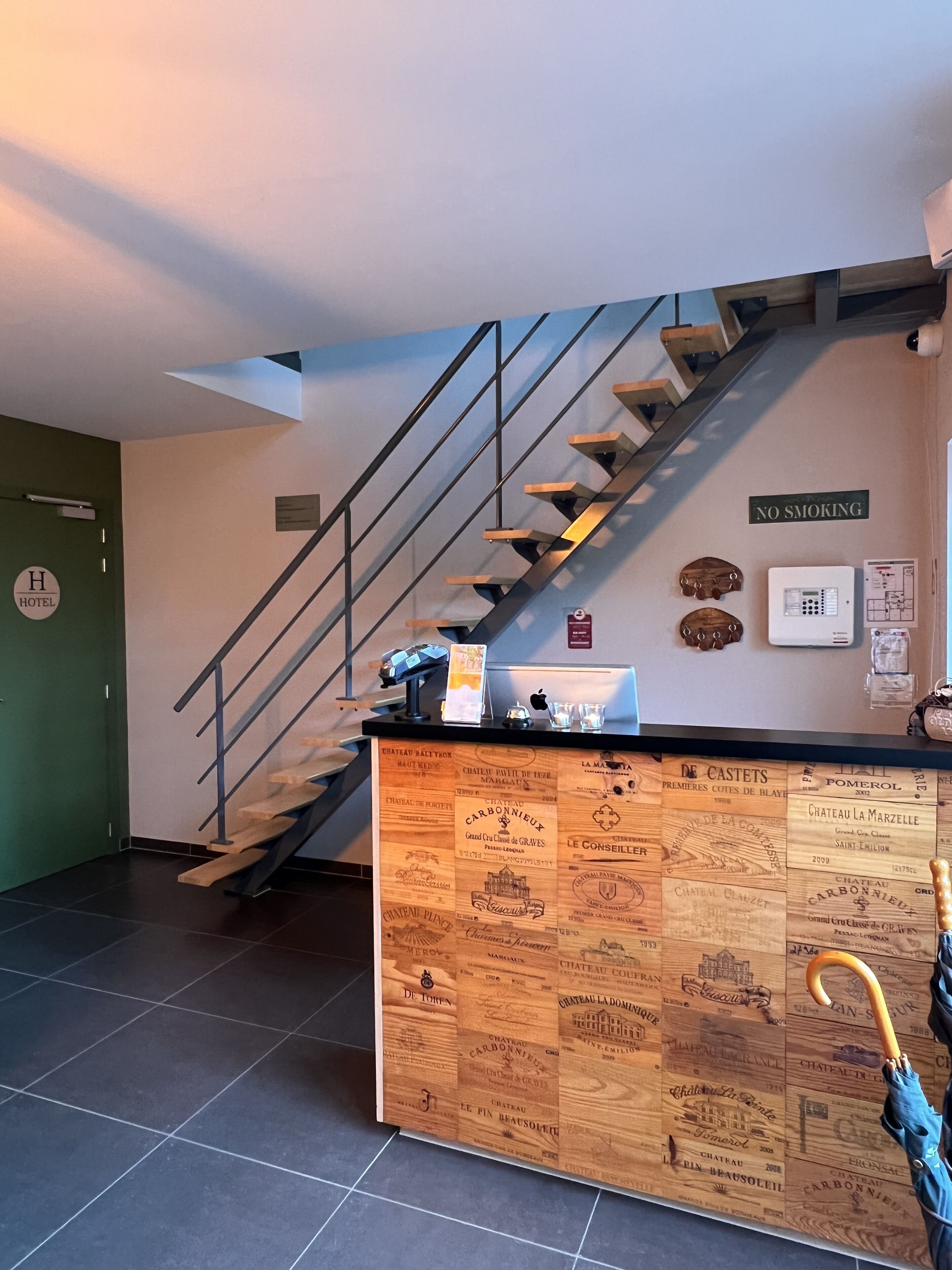 A hotel reception area with a wooden counter displaying wine labels, a staircase, and a 'No Smoking' sign.
