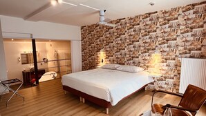 A bedroom with a bed, a wooden floor, a mirror, a chair, and a wall covered with photos. - Hotel Het Heilig Genot (Vleteren)