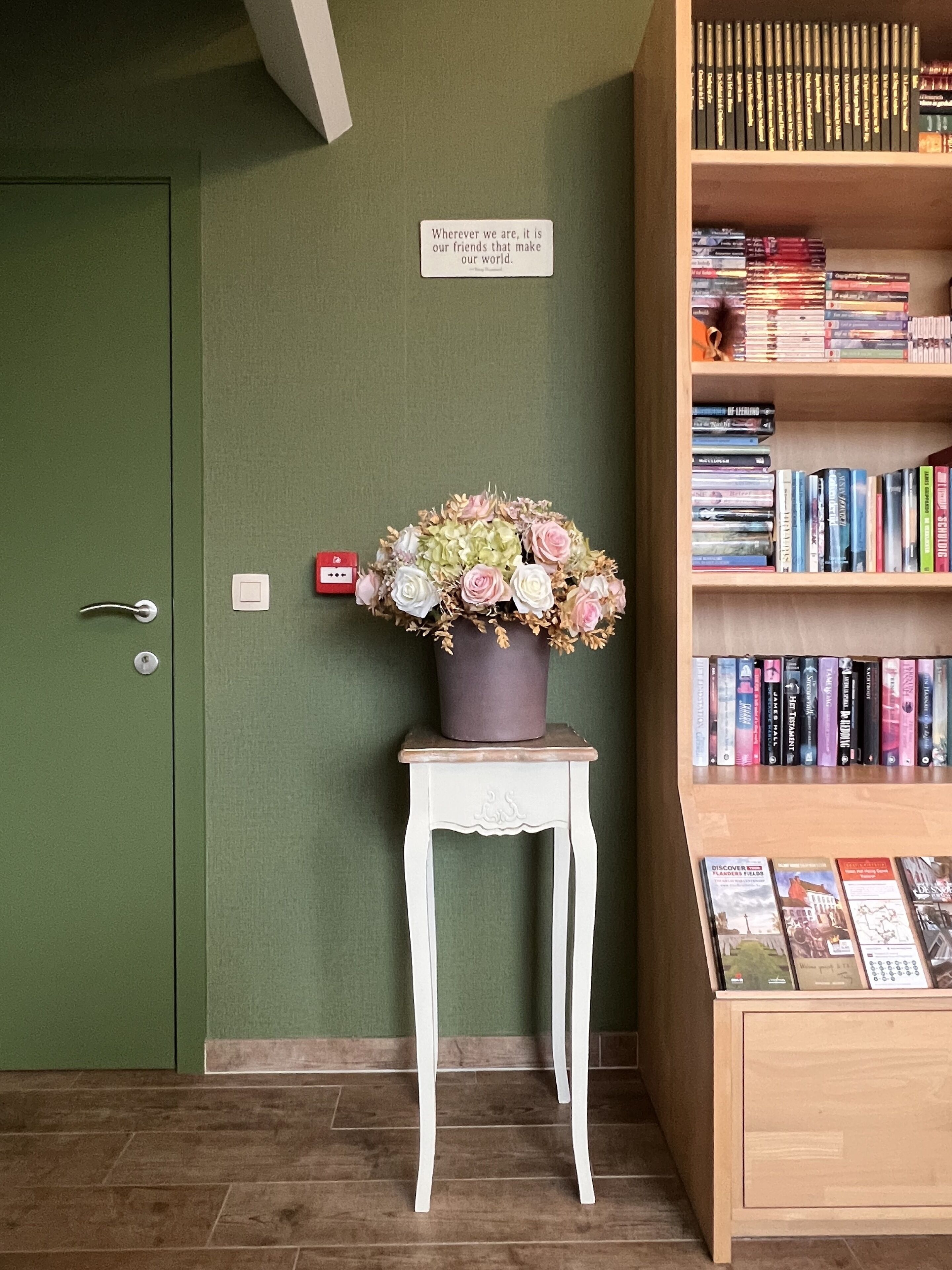 Una puerta verde, un estante blanco con libros y una pequeña mesa con un arreglo floral.