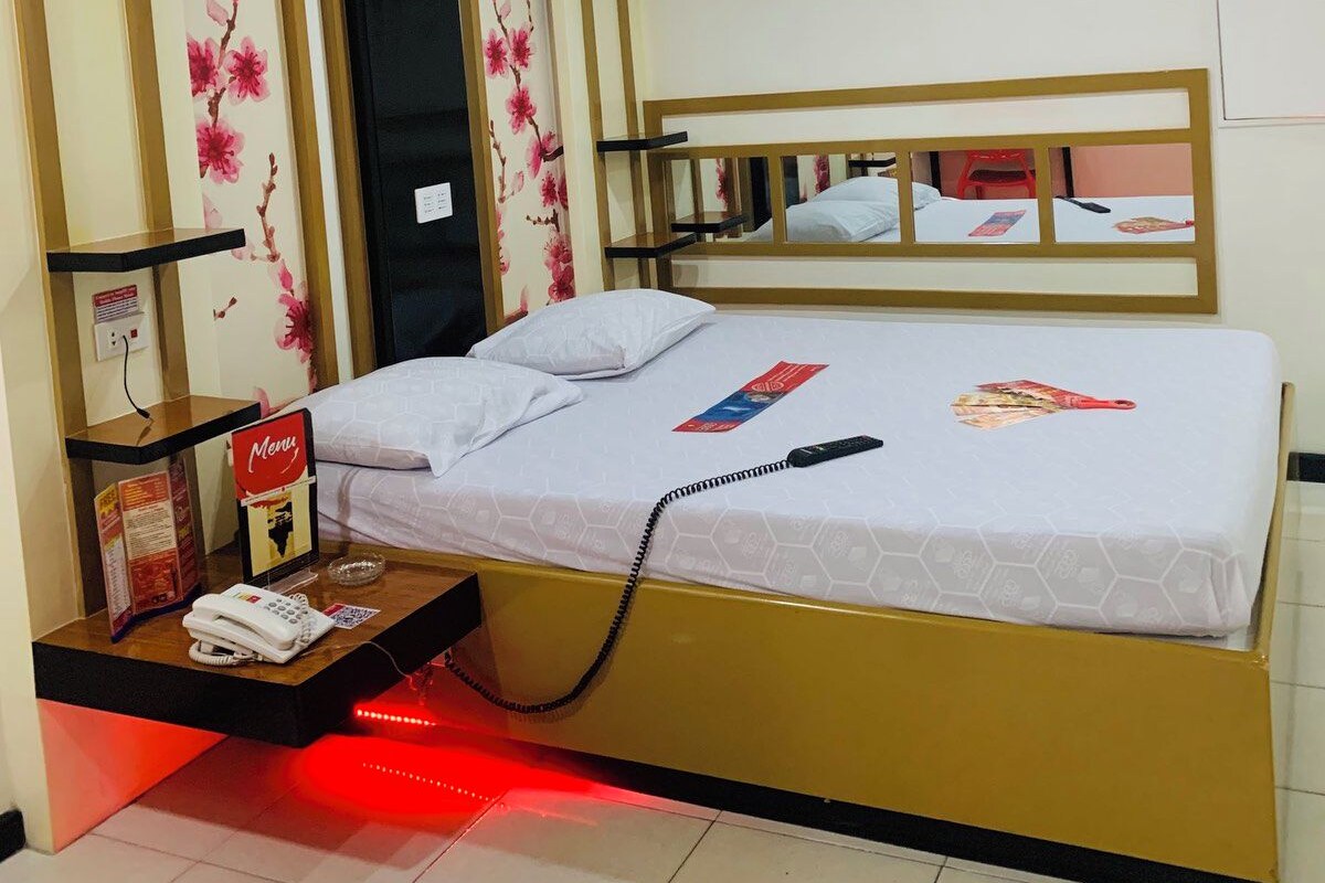 A hotel room with a bed, a bedside table with a phone, a red light, and floral wallpaper.