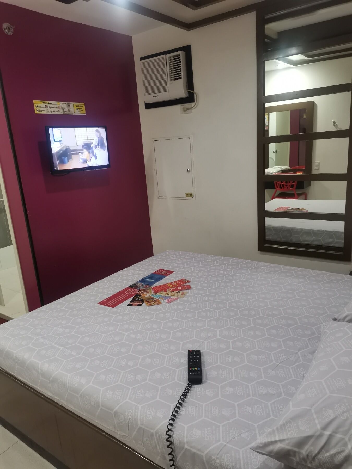 Una habitación de hotel con cama, televisor montado en la pared, una puerta roja y ventana con vistas al exterior.