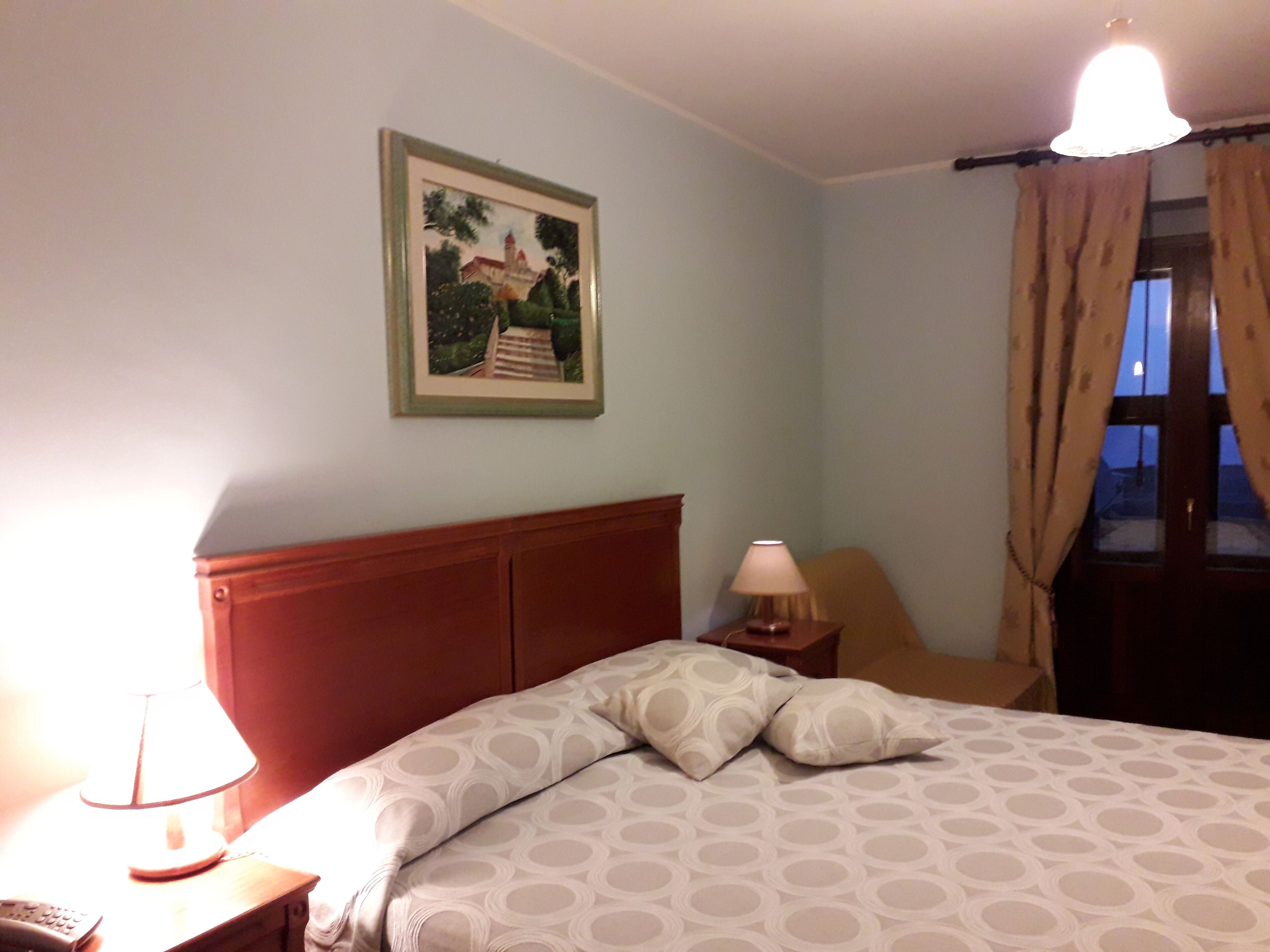 Ein Hotelzimmer mit einem Bett, einem Nachttisch, einer Lampe, einem Bild an der Wand und einem Fenster mit Vorhängen.