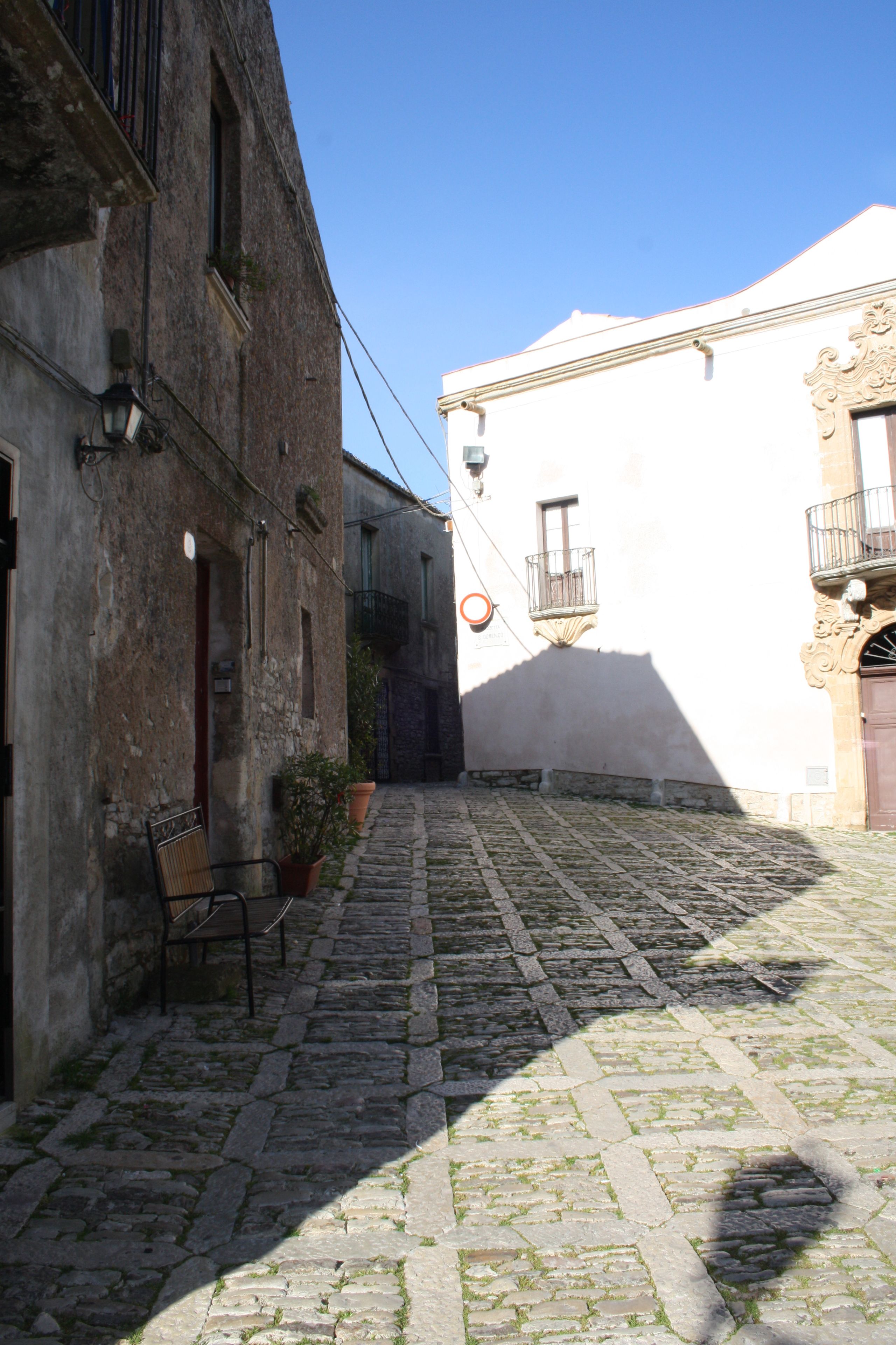 Una estrecha calle adoquinada entre edificios con paredes de piedra, un banco y macetas con plantas.