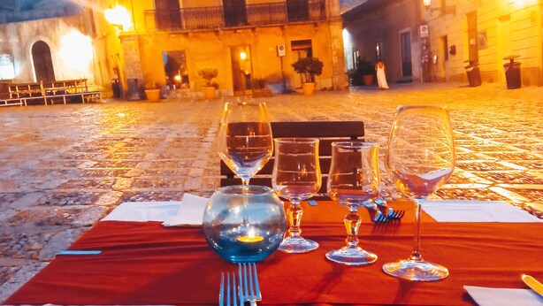 A table set for two with wine glasses and cutlery, a red tablecloth, and a cobblestone street in the background. - Hotel Edelweiss (Erice)