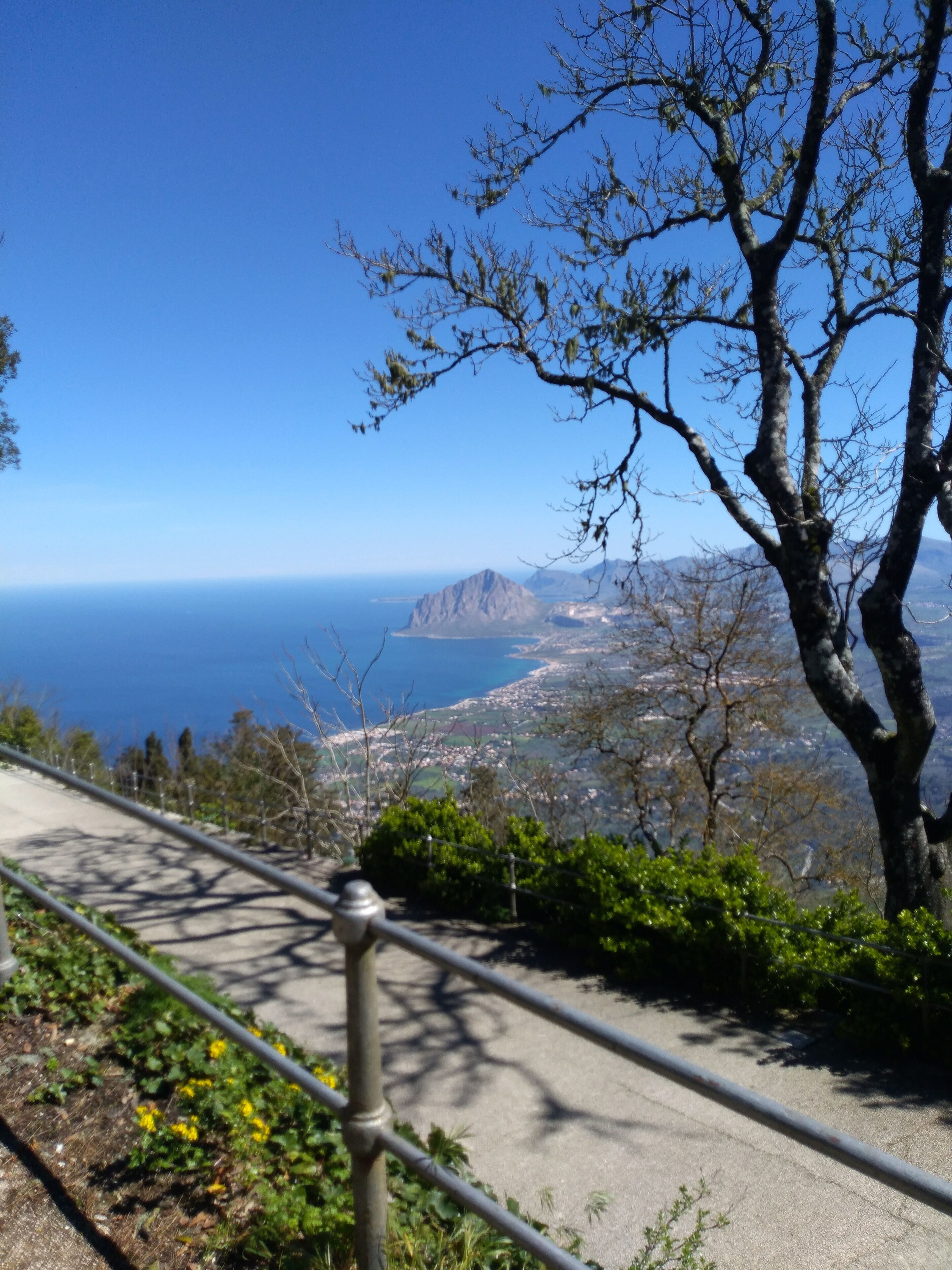 A pathway with a metal railing, surrounded by greenery and a clear view of a coastal landscape with mountains and the sea.