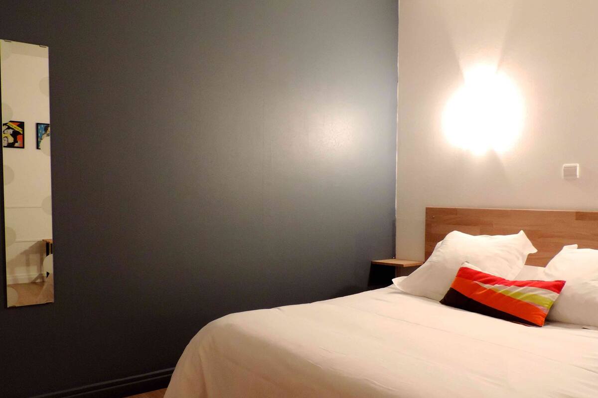 Quarto de hotel com cama, mesa de cabeceira, luminária na parede e espelho.