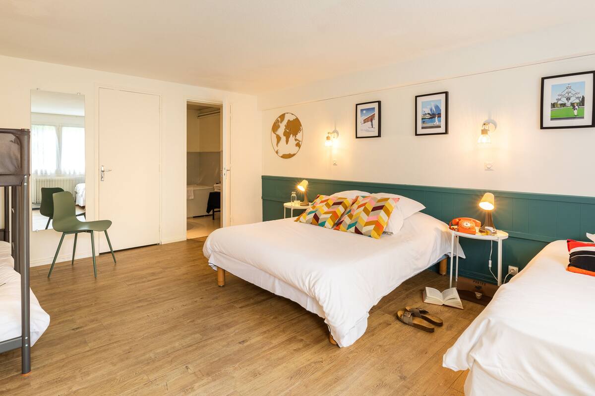 Quarto de hotel com cama de beliche, uma cama de solteiro, uma escrivaninha com luminária e uma cadeira.
