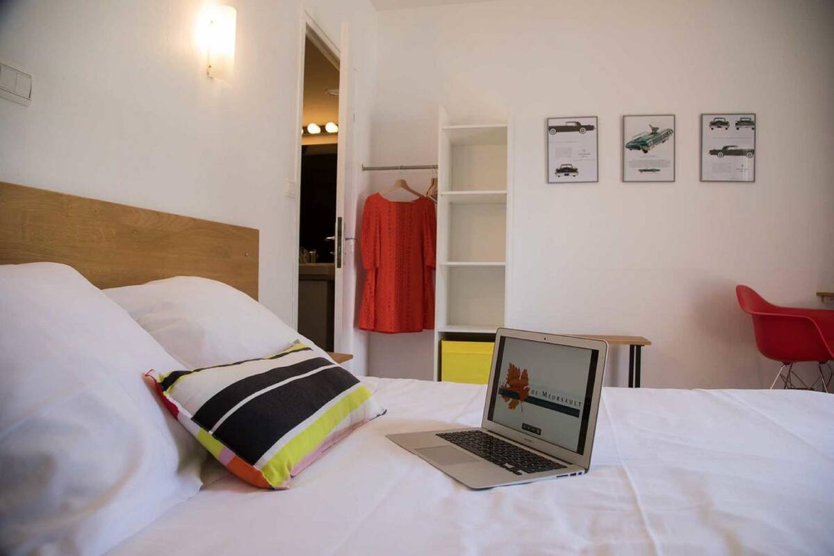 Quarto de hotel com uma cama, um laptop, uma cadeira vermelha e um guarda-roupa com roupas.