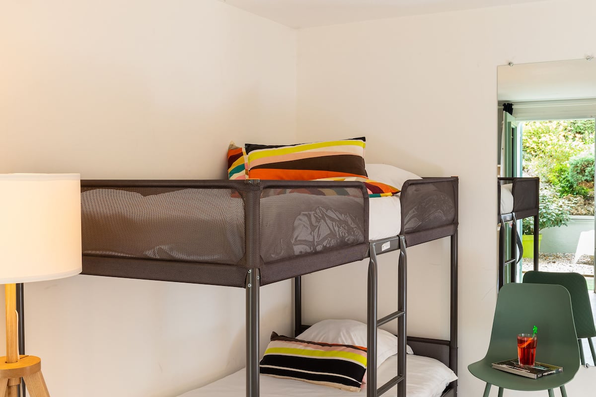 Uma cama beliche com escrivaninha e cadeira, uma luminária e uma janela com vista para o verde.