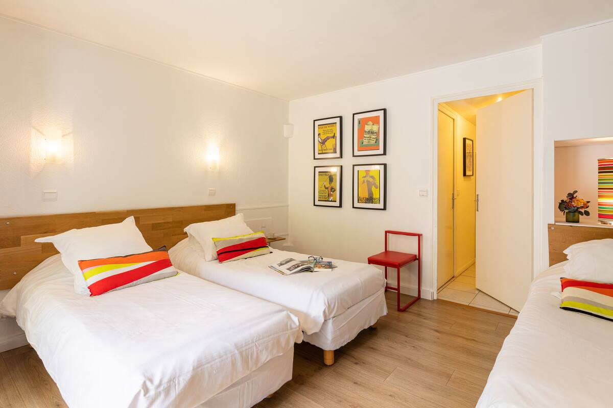 Quarto de hotel com duas camas de solteiro, uma pequena mesa e uma porta que dá para outro quarto.