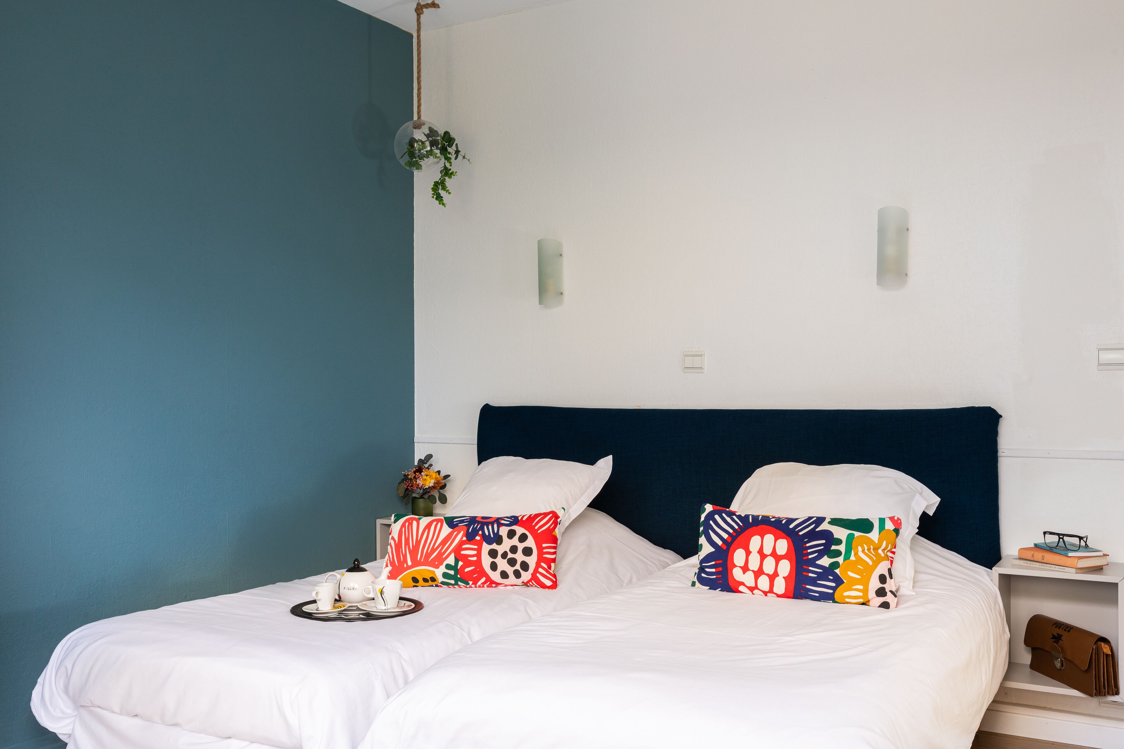 A double bed with white bedding and two colorful pillows, a bedside table with a vase of flowers, and a small shelf with a book and glasses.