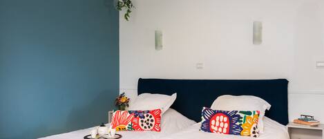 A double bed with white bedding and two colorful pillows, a bedside table with a vase of flowers, and a small shelf with a book and glasses.