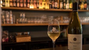 A bottle of Meursault wine and a glass of white wine on a bar counter with shelves of various bottles in the background. - Hôtel Les Hauts de Meursault (Meursault)
