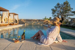 Une femme se détend au bord d’une piscine, un verre et des en-cas à portée de main, avec un kiosque et un mur de pierre en arrière-plan.