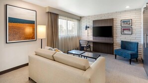 A modern hotel room with a beige sofa, a desk, a chair, a flat-screen TV mounted on a brick wall, and a large framed artwork on the wall. - The Gibson Hotel Great Falls, an Ascend Collection Hotel (Great Falls)