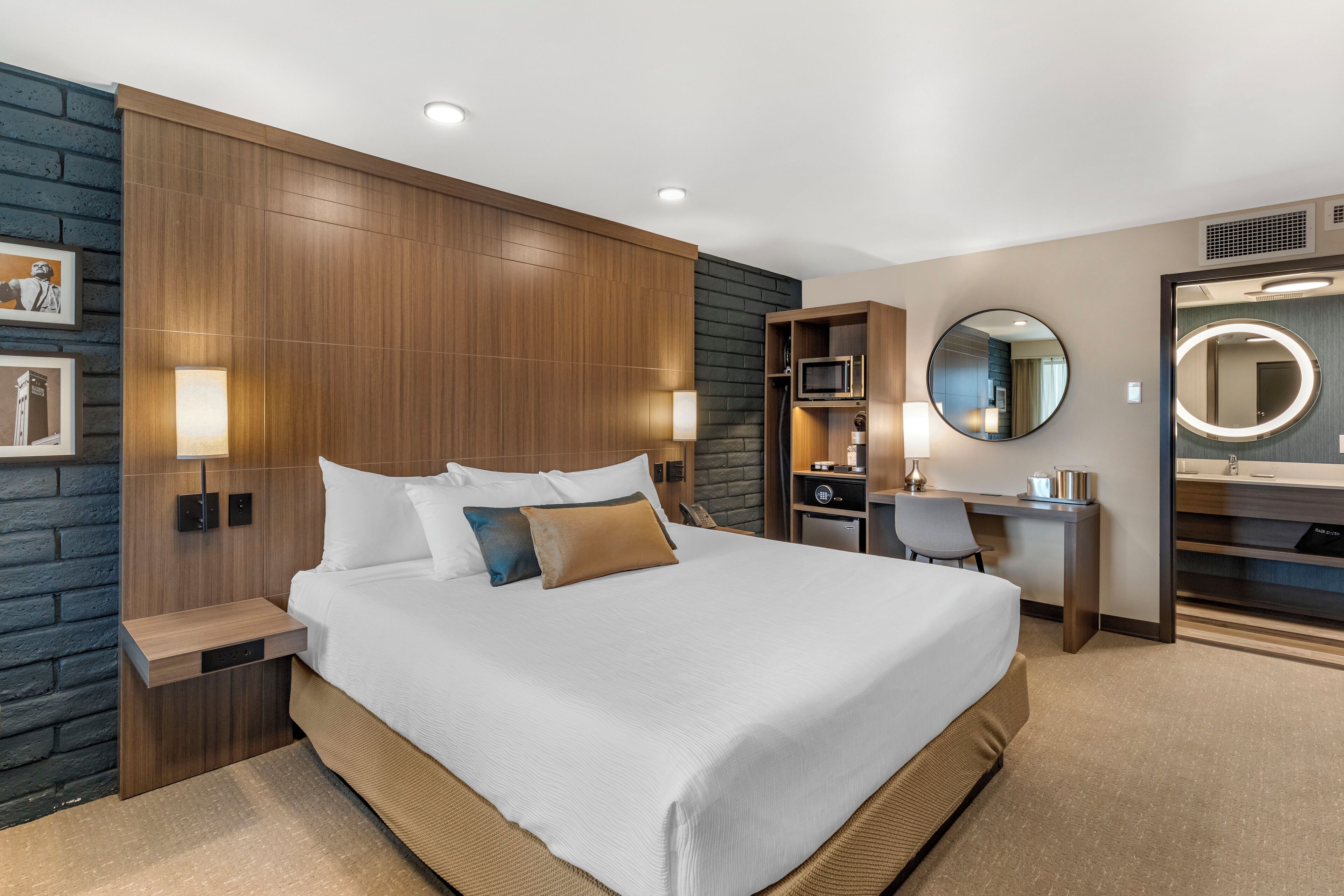 A modern hotel room with a large bed, a desk, a chair, a round mirror, and a small kitchenette.