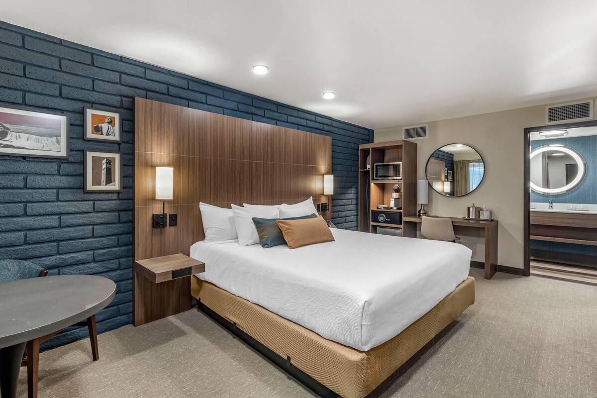 A modern hotel room with a large bed, a wooden headboard, a round mirror, a small desk, and a round table.