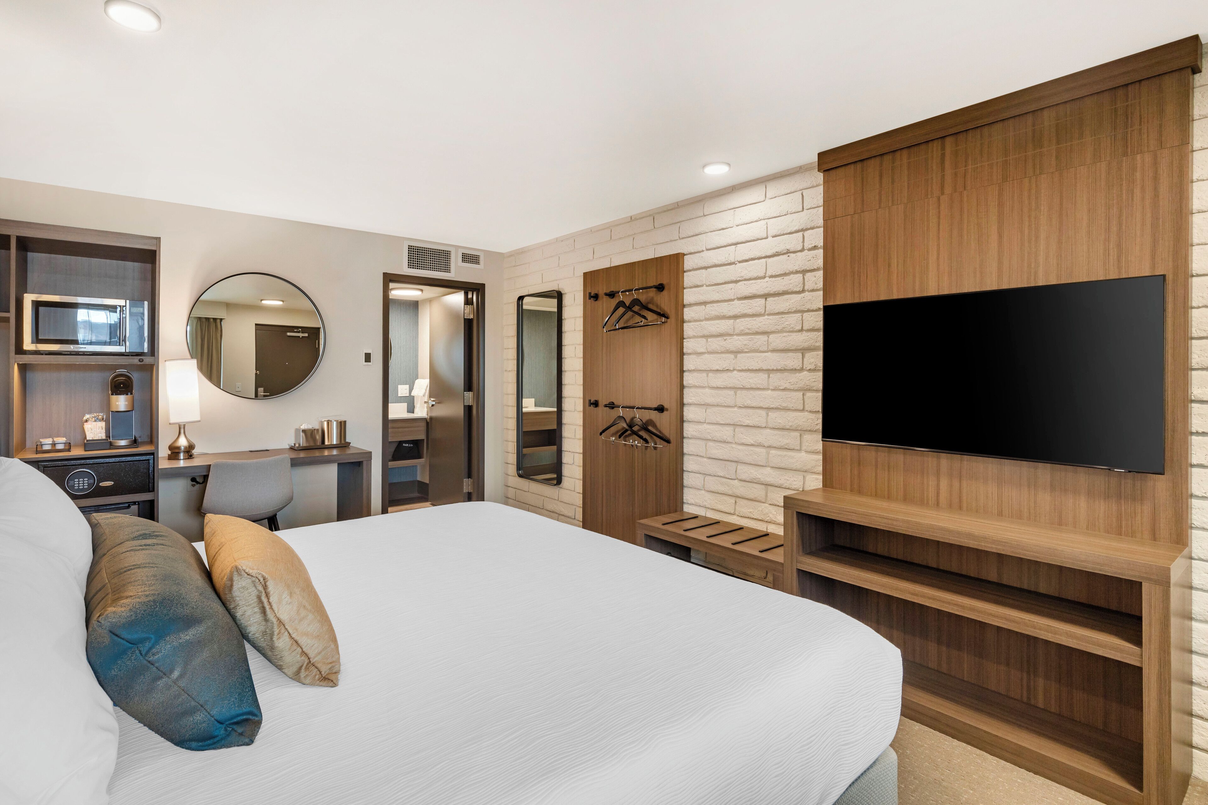 A modern hotel room with a large bed, a flat-screen TV mounted on a wooden panel, a desk with a lamp, a mirror, and a wardrobe.