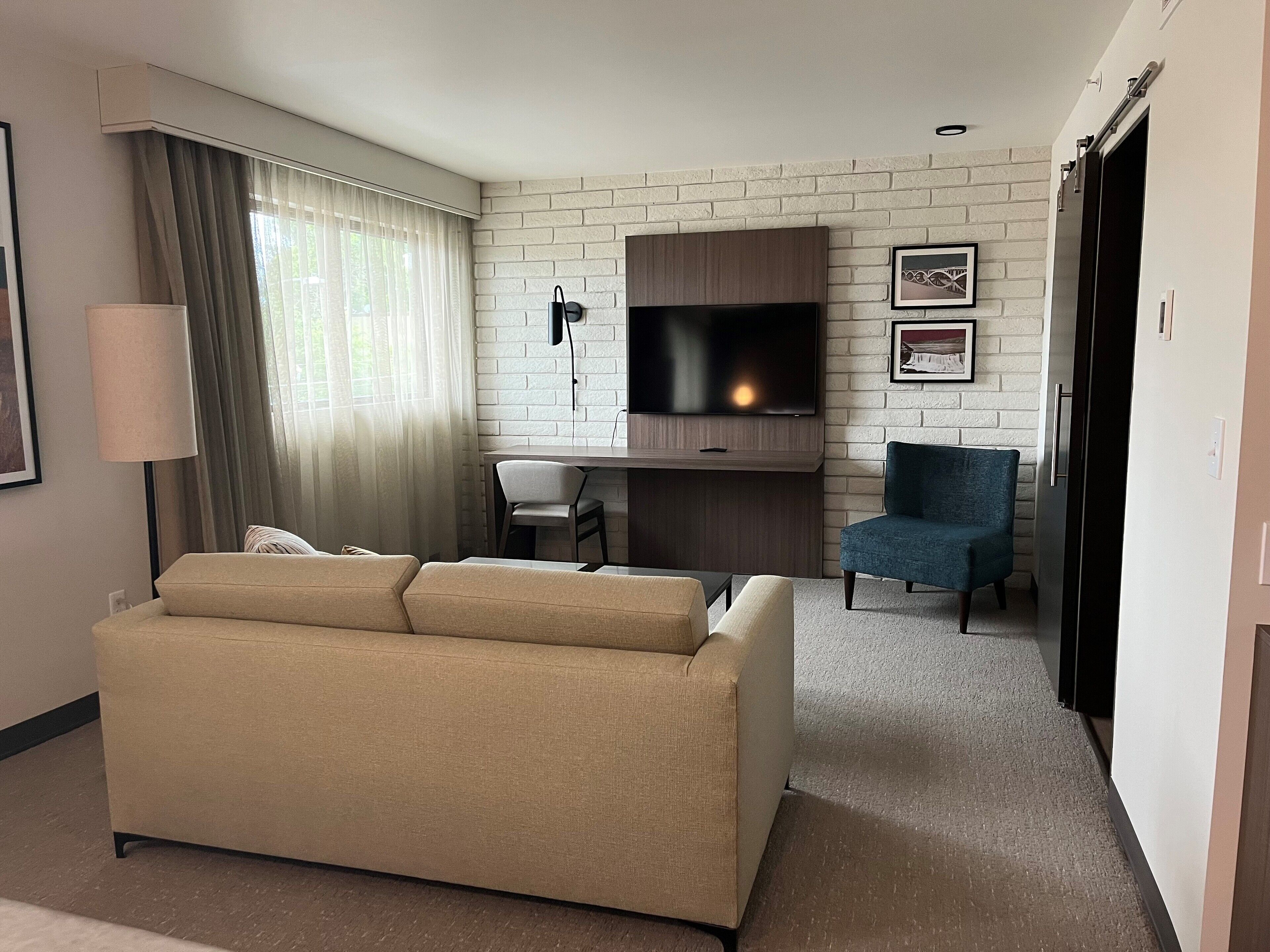 Una habitación de hotel moderna con un sofá color beige, un escritorio con una silla, un televisor y un cuadro en la pared.