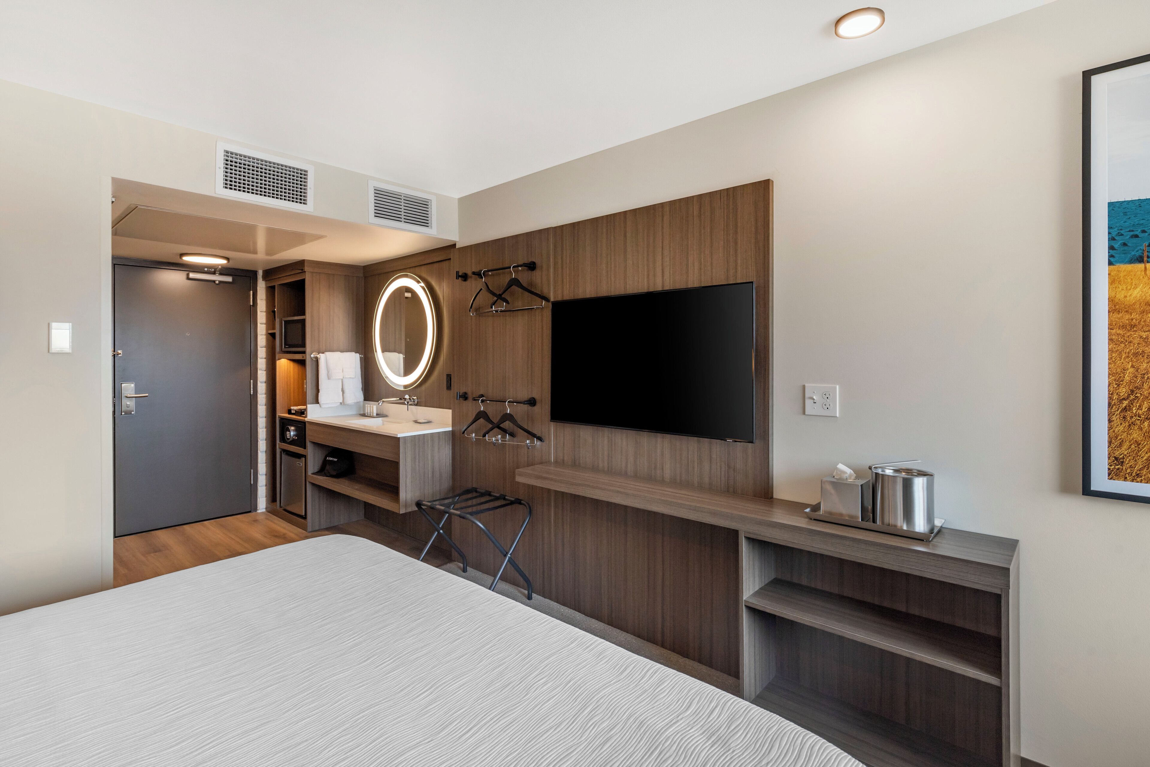 Ein modernes Hotelzimmer mit einem großen Bett, einem an der Wand montierten Fernseher, einem Schminktisch mit Spiegel und einer kleinen Küchenzeile.