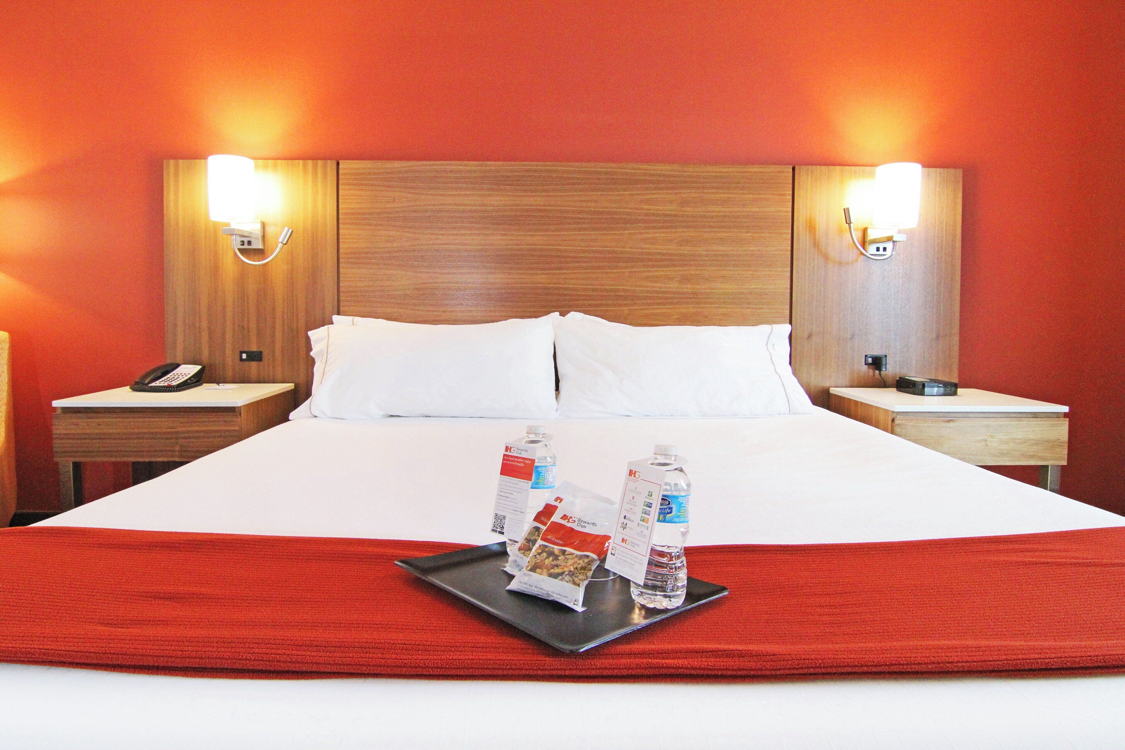 A hotel room with a large bed, two bedside tables, a nightstand, a tray with bottled water and snacks, and two wall-mounted lamps.