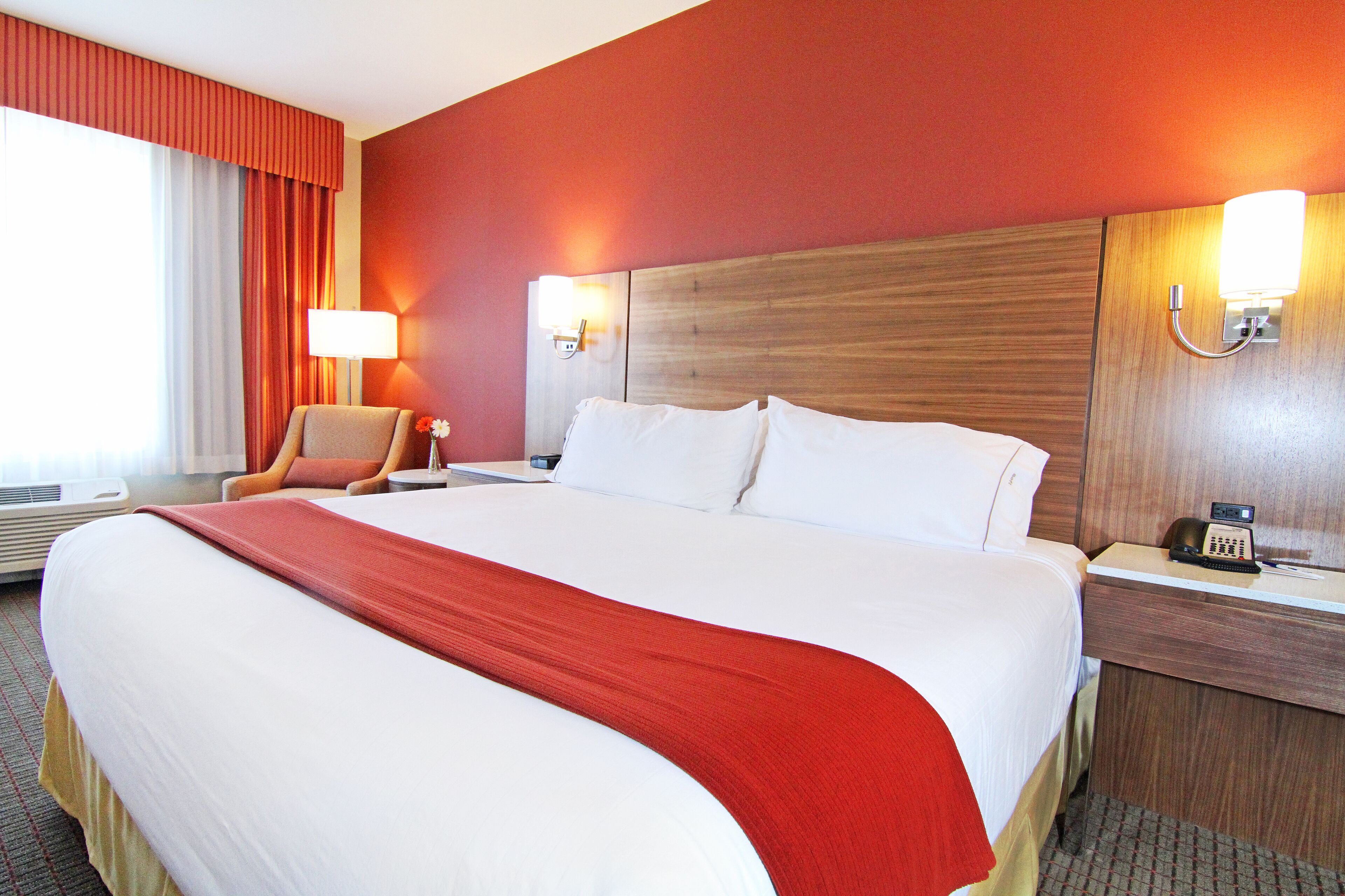 A hotel room with a large bed, a chair, a nightstand with a lamp, and a wall-mounted phone.