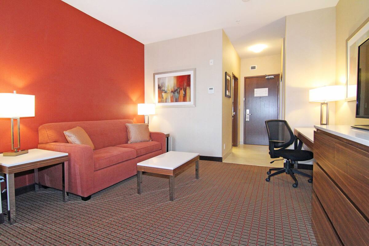 A hotel room with a red wall, a sofa, a coffee table, a desk, a chair, and a bed.