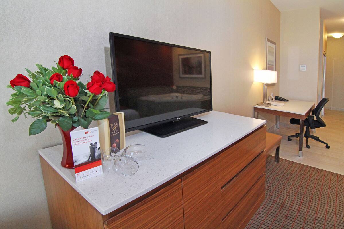 A hotel room with a TV on a white desk, a vase of red roses, a remote control, and a chair.