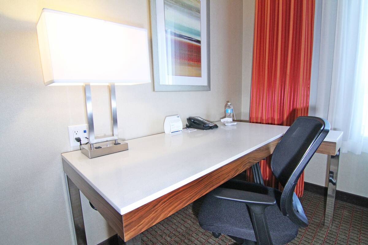 A hotel room with a desk, a lamp, a telephone, a water bottle, and a chair.