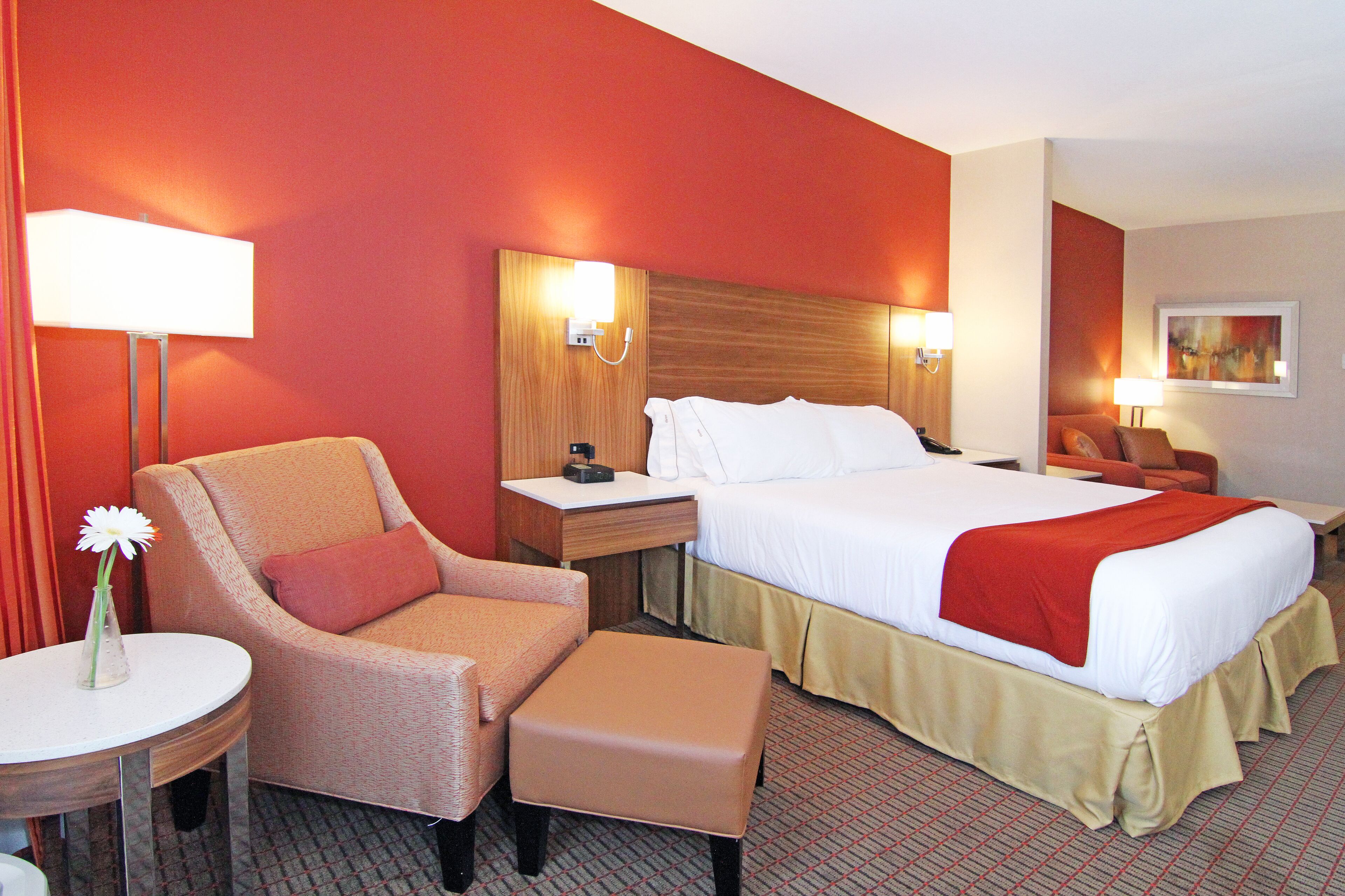 A hotel room with a bed, a chair, a bedside table, a lamp, and a painting on the wall.