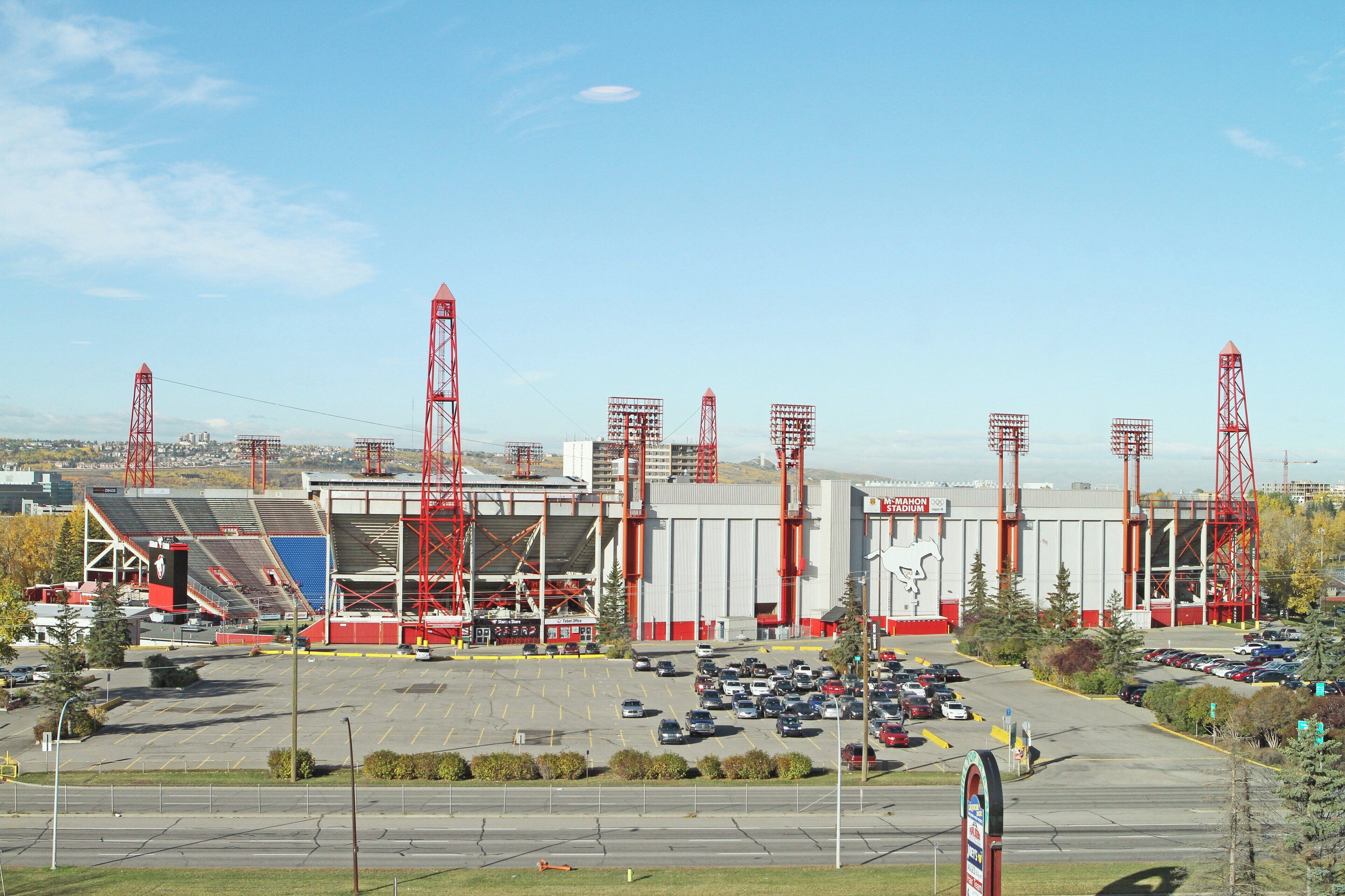 Um estádio com elementos estruturais vermelhos, um estacionamento com carros e um céu claro.