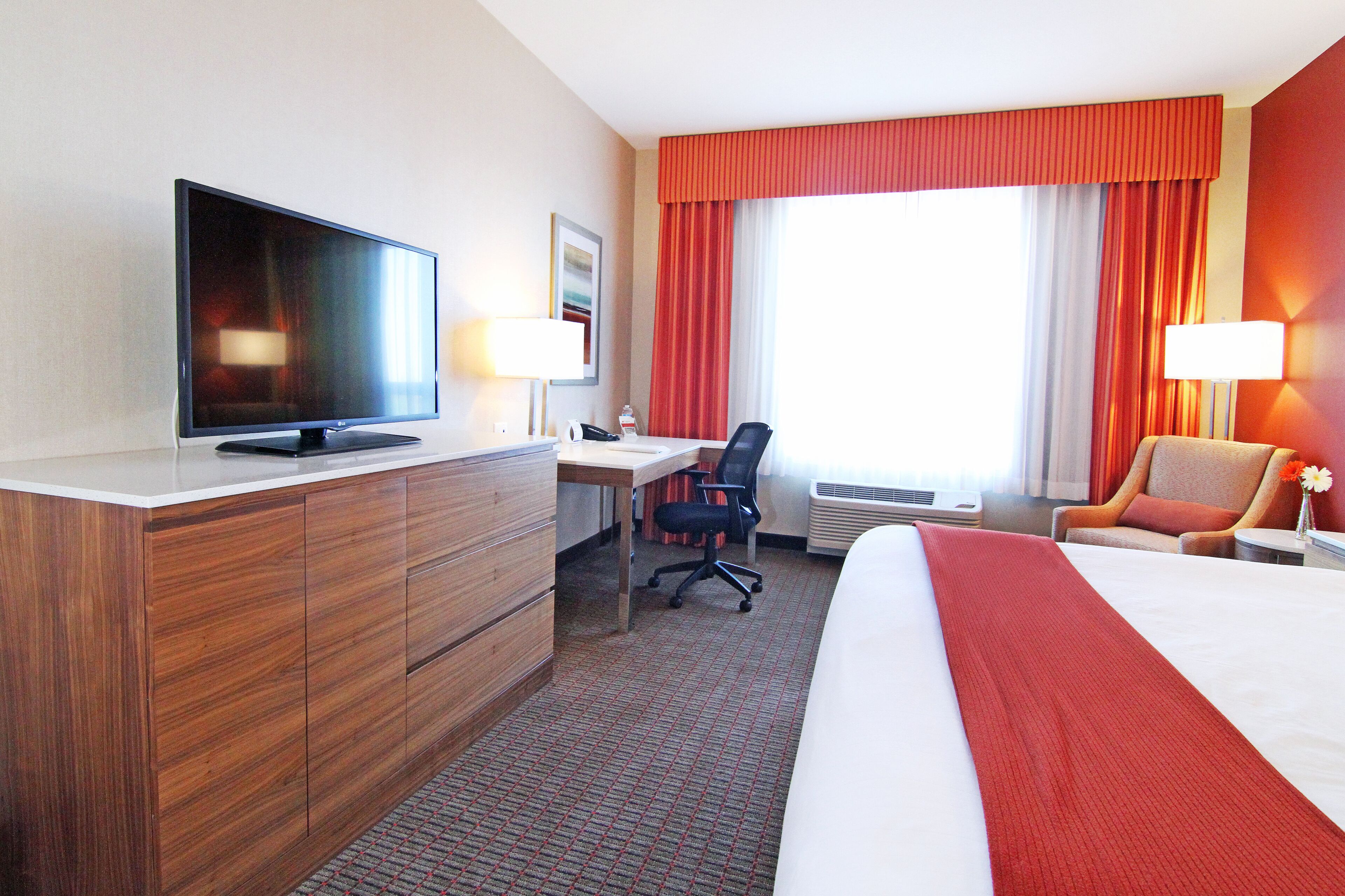 A hotel room with a bed, a desk with a computer, a television, a chair, and a window with curtains.