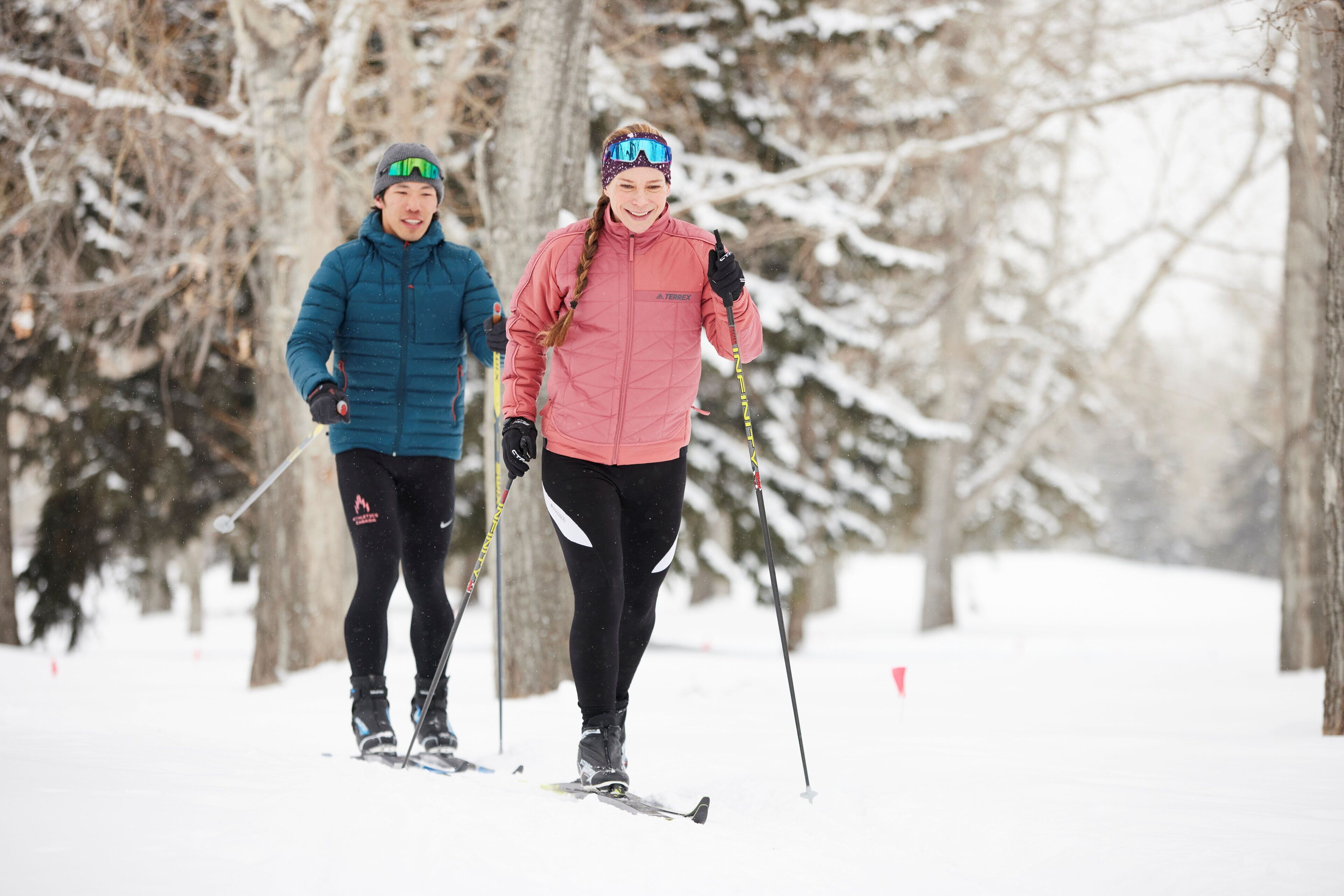 Two people cross-country skiing on a snowy path with trees in the background.