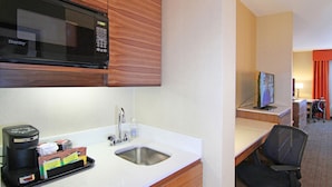 A hotel room with a kitchenette, including a microwave, coffee maker, and sink. There is a desk with a computer and a chair.