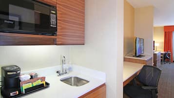 A hotel room with a kitchenette, including a microwave, coffee maker, and sink. There is a desk with a computer and a chair.